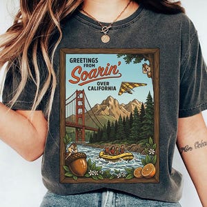 Puede incluir: Camiseta gris oscuro con un dise&ntilde;o enmarcado. El dise&ntilde;o muestra el texto "Greetings from Soarin' Over California" y representa el Golden Gate Bridge, monta&ntilde;as, un ala delta y una escena de rafting con Chip y Chop.