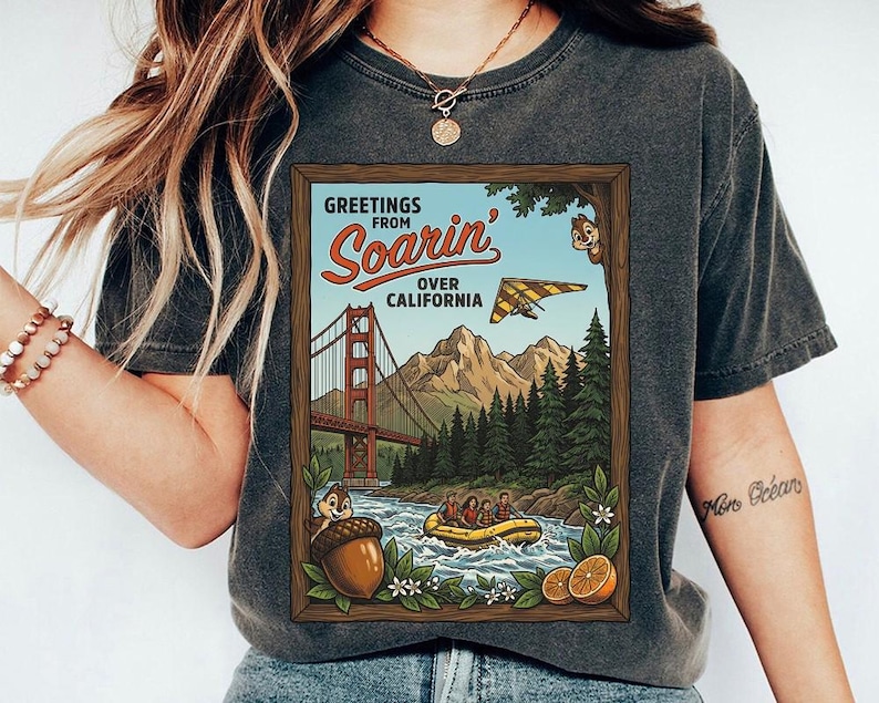 Puede incluir: Camiseta gris oscuro con un dise&ntilde;o enmarcado. El dise&ntilde;o muestra el texto "Greetings from Soarin' Over California" y representa el Golden Gate Bridge, monta&ntilde;as, un ala delta y una escena de rafting con Chip y Chop.