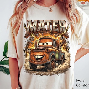 May include: Ivory Comfort Color t-shirt featuring a cartoon graphic of Mater from the movie Cars. The graphic includes the word "MATER" in a distressed, 3D font above the character. Mater is depicted as a rusty tow truck with a smiling face.
