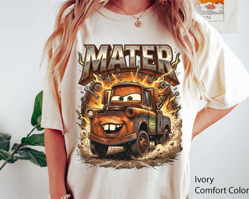 May include: Ivory Comfort Color t-shirt featuring a cartoon graphic of Mater from the movie Cars. The graphic includes the word "MATER" in a distressed, 3D font above the character. Mater is depicted as a rusty tow truck with a smiling face.