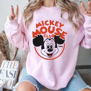 May include: A light pink sweatshirt featuring the text "Mickey Mouse Club" in red and black, with a graphic of Mickey Mouse. The sweatshirt has long sleeves and a crew neck.