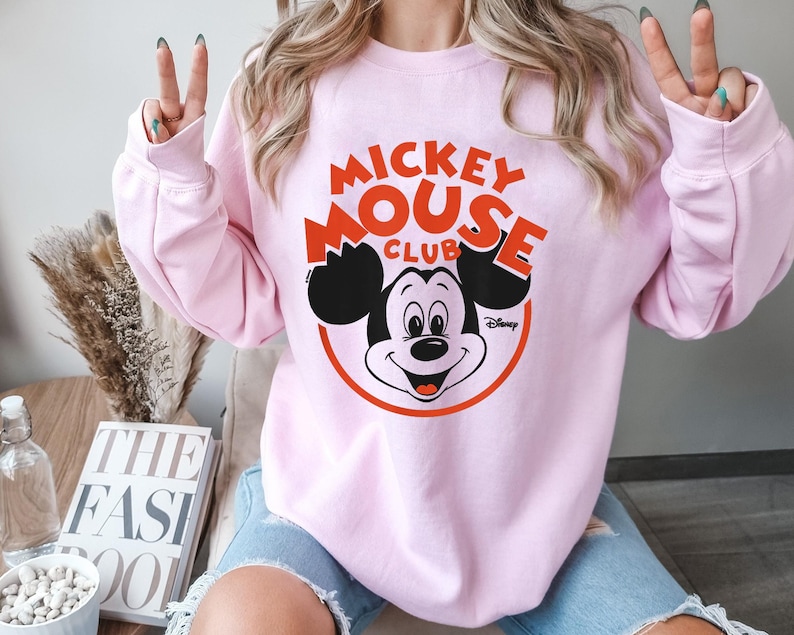 May include: A light pink sweatshirt featuring the text "Mickey Mouse Club" in red and black, with a graphic of Mickey Mouse. The sweatshirt has long sleeves and a crew neck.