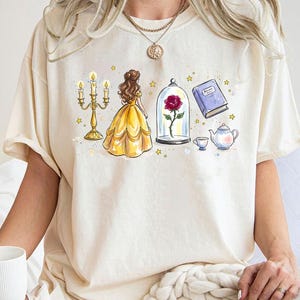 May include: Cream-colored t-shirt featuring a watercolor illustration of a yellow gown, a candelabra, a rose in a glass dome, a book, and a teapot. The design includes gold stars and a teacup.