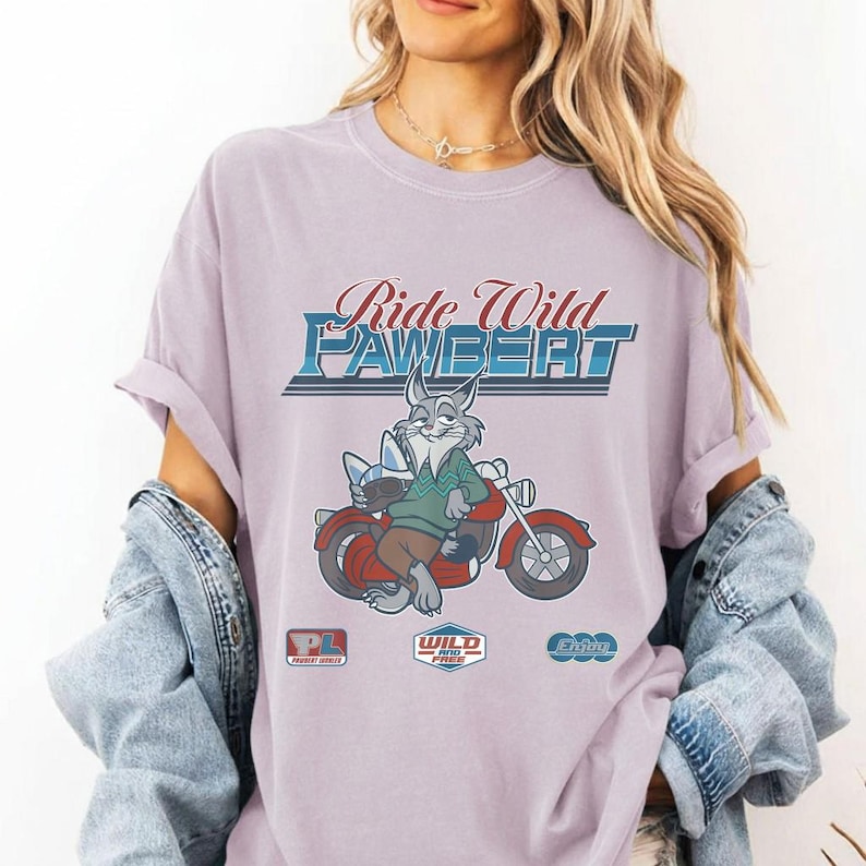 Puede incluir: Una camiseta de color lila con el texto "Ride Wild PAWBERT" sobre una ilustraci&oacute;n de un conejo montando una moto roja. El conejo lleva un su&eacute;ter verde y pantalones marrones. La camiseta tambi&eacute;n tiene tres logotipos peque&ntilde;os.