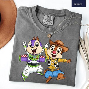 May include: A gray Comfort Colors t-shirt featuring a cartoon graphic of two animated chipmunks. One is dressed as Buzz Lightyear, the other as a cowboy. The shirt has a crew neck and short sleeves. A silver necklace with a pendant is draped across the shirt.