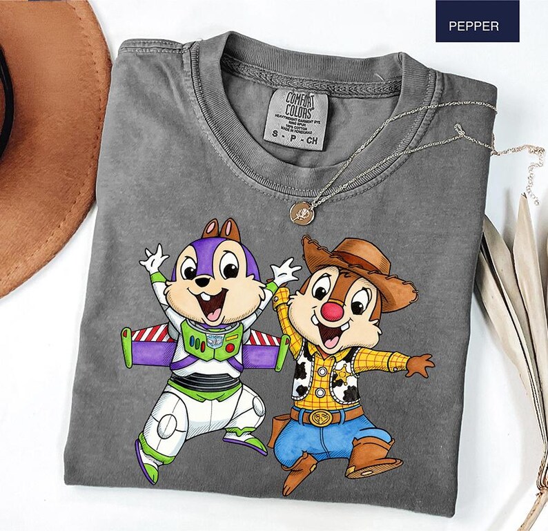 May include: A gray Comfort Colors t-shirt featuring a cartoon graphic of two animated chipmunks. One is dressed as Buzz Lightyear, the other as a cowboy. The shirt has a crew neck and short sleeves. A silver necklace with a pendant is draped across the shirt.