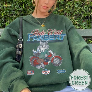 Puede incluir: Sudadera verde bosque con el texto "Ride Wild PAWBERT" y un conejo de dibujos animados montado en una moto roja. La sudadera tiene tres peque&ntilde;os parches y una etiqueta redonda que dice "Forest Green".
