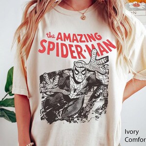 May include: An ivory short-sleeve t-shirt featuring a black and white graphic of the comic book character Spider-Man in a dynamic pose. Above the graphic, the text reads "the AMAZING SPIDER-MAN" in bold red capital letters. The shirt has a crew neckline.