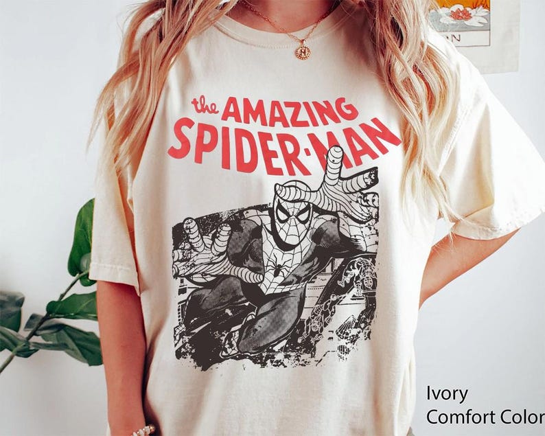 May include: An ivory short-sleeve t-shirt featuring a black and white graphic of the comic book character Spider-Man in a dynamic pose. Above the graphic, the text reads "the AMAZING SPIDER-MAN" in bold red capital letters. The shirt has a crew neckline.