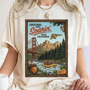 Puede incluir: Camiseta color crema con un dise&ntilde;o enmarcado. El dise&ntilde;o muestra "Greetings from Soarin' Over California" y representa el Golden Gate Bridge, monta&ntilde;as, un ala delta y un r&iacute;o con una balsa. El dise&ntilde;o incluye personajes de dibujos animados y elementos florales.