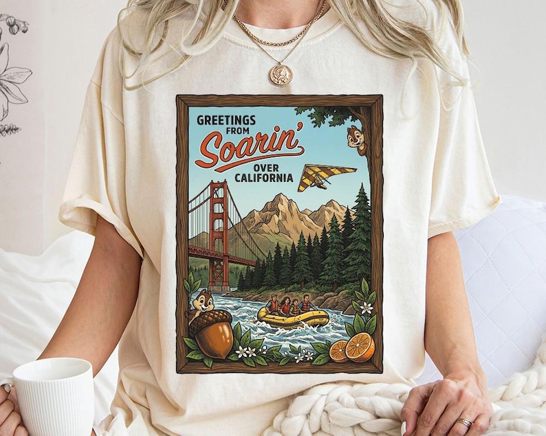 Puede incluir: Camiseta color crema con un dise&ntilde;o enmarcado. El dise&ntilde;o muestra "Greetings from Soarin' Over California" y representa el Golden Gate Bridge, monta&ntilde;as, un ala delta y un r&iacute;o con una balsa. El dise&ntilde;o incluye personajes de dibujos animados y elementos florales.