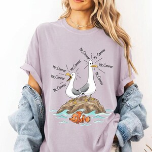 May include: A lilac-colored t-shirt featuring a cartoon illustration of two seagulls on a rock, with the text "Mr. Connor" emanating from them. Below, a cartoon clownfish swims in blue water. The shirt is part of a clothing collection.