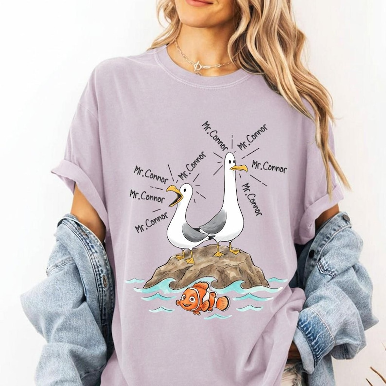 May include: A lilac-colored t-shirt featuring a cartoon illustration of two seagulls on a rock, with the text "Mr. Connor" emanating from them. Below, a cartoon clownfish swims in blue water. The shirt is part of a clothing collection.