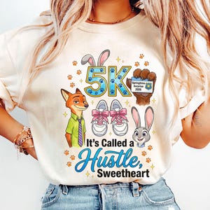 May include: Cream-coloured t-shirt with cartoon characters and the text "5K". The design includes a fox in a green shirt and purple tie, a bunny, running shoes with pink laces, and the phrase "It's Called a Hustle, Sweetheart".