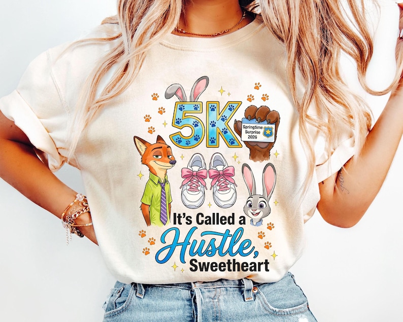 May include: Cream-coloured t-shirt with cartoon characters and the text "5K". The design includes a fox in a green shirt and purple tie, a bunny, running shoes with pink laces, and the phrase "It's Called a Hustle, Sweetheart".