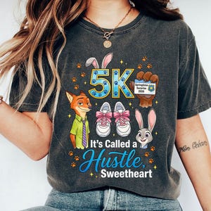May include: Dark grey t-shirt with cartoon characters and the text "5K". The design includes a fox, a rabbit, and running shoes with pink laces. The text "It's Called a Hustle, Sweetheart" is also displayed.