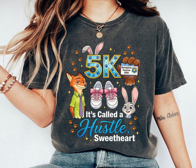 May include: Dark grey t-shirt with cartoon characters and the text "5K". The design includes a fox, a rabbit, and running shoes with pink laces. The text "It's Called a Hustle, Sweetheart" is also displayed.