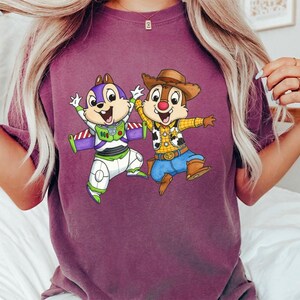 May include: A maroon t-shirt featuring a cartoon graphic of two animated chipmunks dressed as Buzz Lightyear and Woody. The Buzz Lightyear chipmunk wears a purple and white space suit. The Woody chipmunk wears a cowboy hat and vest.