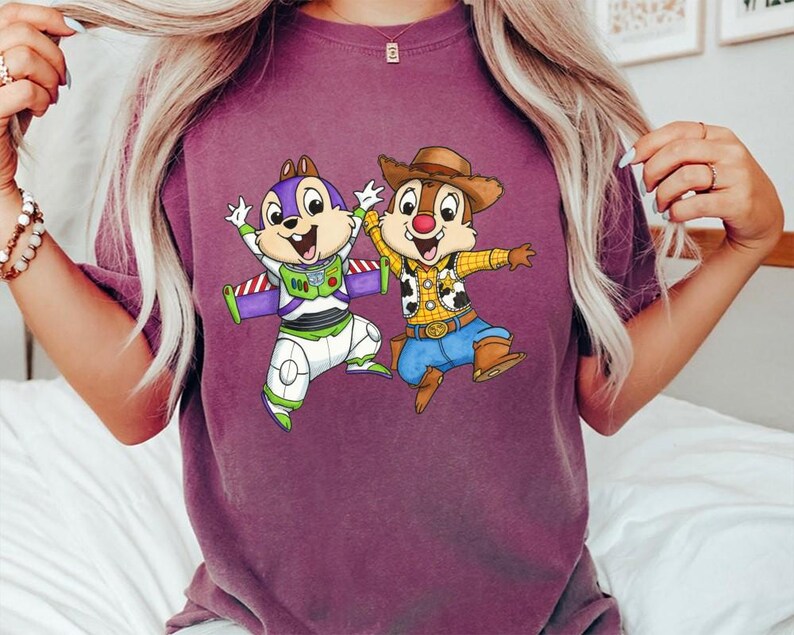 May include: A maroon t-shirt featuring a cartoon graphic of two animated chipmunks dressed as Buzz Lightyear and Woody. The Buzz Lightyear chipmunk wears a purple and white space suit. The Woody chipmunk wears a cowboy hat and vest.