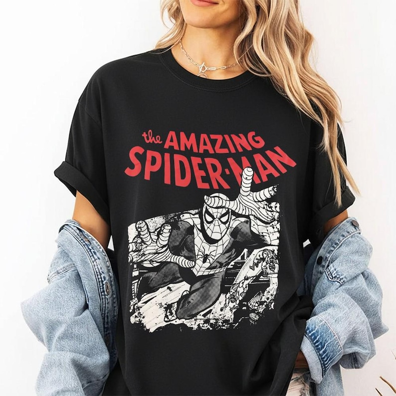 May include: A black short-sleeve t-shirt featuring a graphic print of the comic book character Spider-Man in a dynamic pose. The text above the illustration reads the AMAZING SPIDER-MAN in bold red letters. The graphic is in a black and white style.