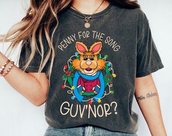 Op de afbeelding: Donkergrijs t-shirt met een cartoon konijn karakter en de tekst "PENNY FOR THE SONG, GUV'NOR?" Het konijn draagt een blauwe sjaal en een rode jas, omringd door kerstverlichting en hulst.
