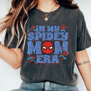 May include: Dark gray t-shirt with the text "IN MY SPIDEY MOM ERA" in blue and red. The design includes a Spider-Man mask and illustrations of Spider-Man.