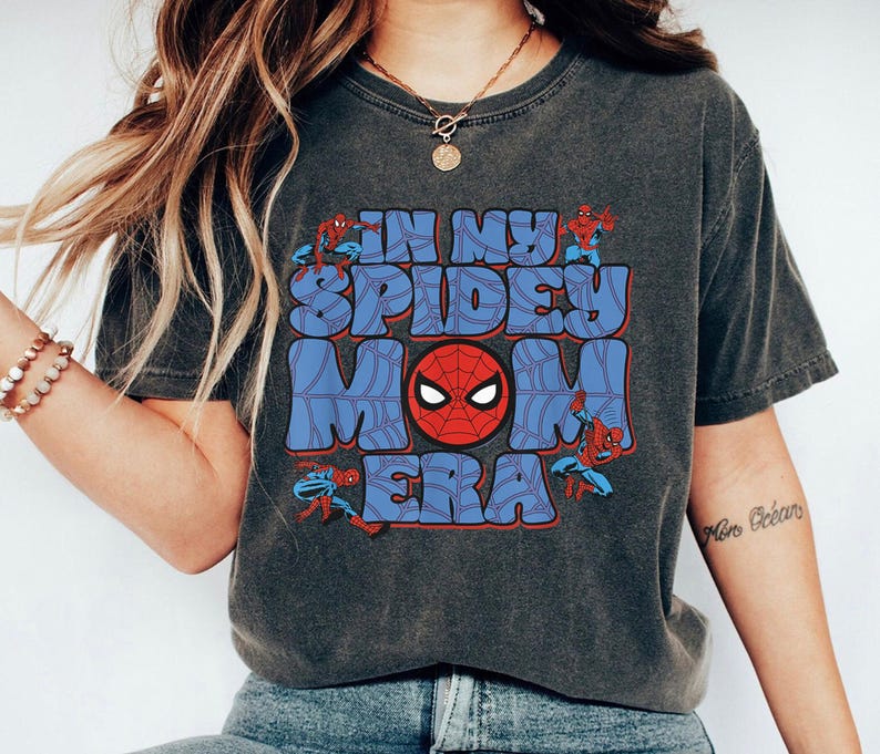 May include: Dark gray t-shirt with the text "IN MY SPIDEY MOM ERA" in blue and red. The design includes a Spider-Man mask and illustrations of Spider-Man.