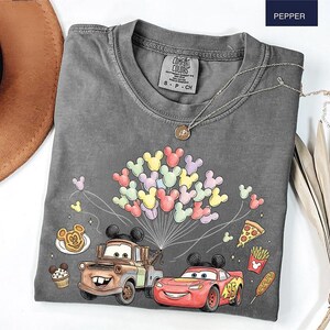 May include: A grey Comfort Colors t-shirt with a graphic of Mickey Mouse balloons, Mater and Lightning McQueen from Cars, and food illustrations. The shirt has a crew neck and short sleeves. A silver necklace with a pendant is also visible.