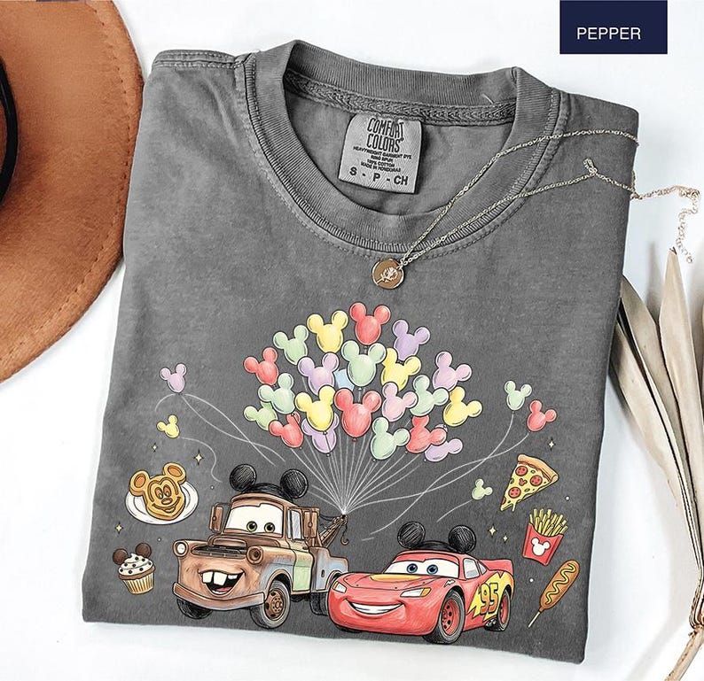 May include: A grey Comfort Colors t-shirt with a graphic of Mickey Mouse balloons, Mater and Lightning McQueen from Cars, and food illustrations. The shirt has a crew neck and short sleeves. A silver necklace with a pendant is also visible.