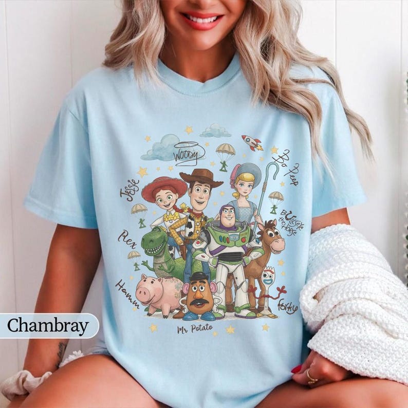May include: A light blue t-shirt featuring a graphic print of Toy Story characters including Woody, Buzz Lightyear, Jessie, Rex, Hamm, Mr. Potato Head, and Forky, surrounded by handwritten-style character names and small star and cloud illustrations.