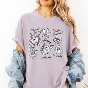 Puede incluir: Una camiseta de color lila claro con ilustraciones de personajes de dibujos animados en blanco y negro, con nombres como "Nick Wilde" y "Dr. Fuzzby". La camiseta tiene cuello redondo y mangas cortas.
