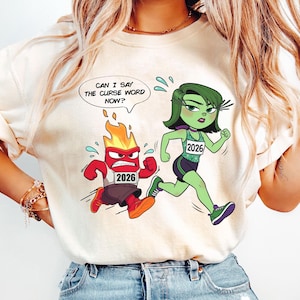 May include: Beige t-shirt featuring a cartoon graphic of two animated characters, one red and one green, in a race. The red character is on fire and the green character is running. Text above the red character reads, "CAN I SAY THE CURSE WORD NOW?"