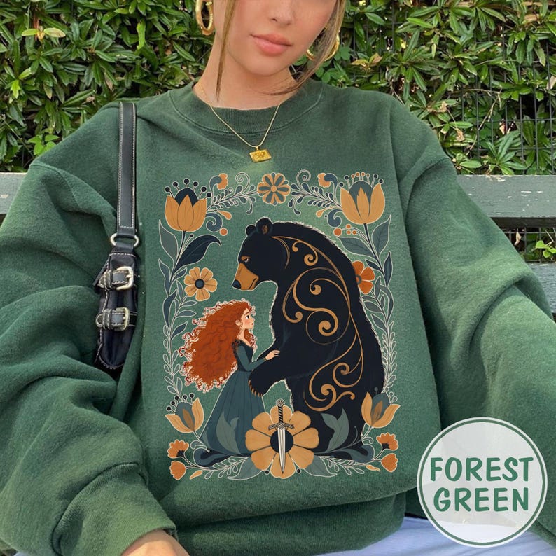 Op de afbeelding: Boszwarte groene sweatshirt met een afbeelding van een beer en een vrouw omringd door bloemen. Het ontwerp bevat een zwaard en de tekst "Forest Green" in een cirkel. De sweatshirt is effen van kleur.