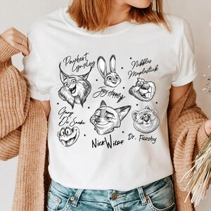 Puede incluir: Camiseta blanca con ilustraciones en blanco y negro de personajes de dibujos animados de animales. La camiseta presenta personajes como un lince, un conejo, un zorro y un oso, cada uno con sus nombres escritos encima o debajo.