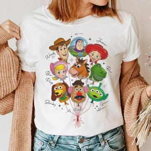 Puede incluir: Camiseta blanca con un gr&aacute;fico colorido de personajes de Toy Story como globos. El dise&ntilde;o incluye a Woody, Buzz Lightyear, Jessie y otros, con sus nombres encima. Un dise&ntilde;o alegre y juguet&oacute;n.