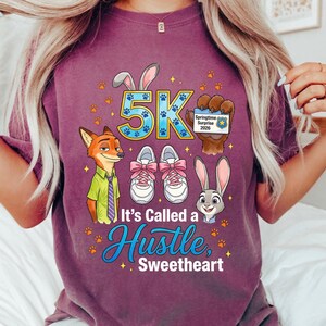 May include: A purple t-shirt with a graphic design. The design includes cartoon characters, the text "5K", and the phrase "It's Called a Hustle, Sweetheart". The design also features paw prints, running shoes, and a Springtime Surprise 2020 badge.