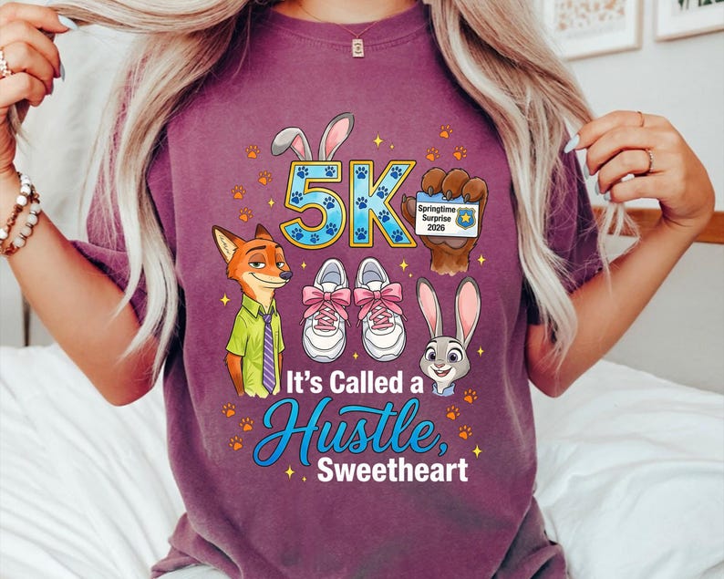 May include: A purple t-shirt with a graphic design. The design includes cartoon characters, the text "5K", and the phrase "It's Called a Hustle, Sweetheart". The design also features paw prints, running shoes, and a Springtime Surprise 2020 badge.