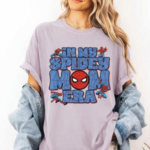May include: A light purple t-shirt with the text "IN MY SPIDEY MOM ERA" in blue bubble letters. The design includes a red Spider-Man mask and several Spider-Man figures in action poses.