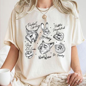 Puede incluir: Camiseta color crema con ilustraciones en blanco y negro de personajes de dibujos animados de animales con sus nombres. Los personajes incluyen un lince, un conejo, un zorro, una rana y un oso. La camiseta tiene un corte holgado.