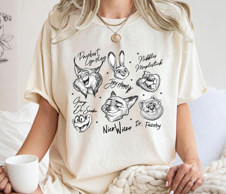 Puede incluir: Camiseta color crema con ilustraciones en blanco y negro de personajes de dibujos animados de animales con sus nombres. Los personajes incluyen un lince, un conejo, un zorro, una rana y un oso. La camiseta tiene un corte holgado.