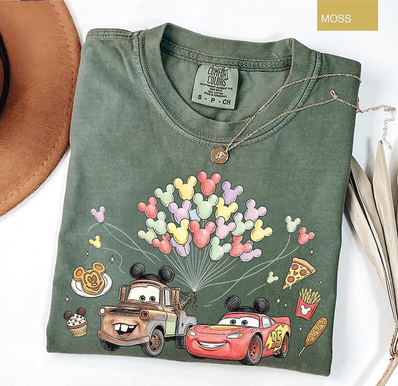 May include: A moss green t-shirt with a graphic of Mickey Mouse balloons, Mater the tow truck, and Lightning McQueen. The shirt also has images of food items like pizza, fries, and a corn dog. The shirt is labelled "Comfort Colors".