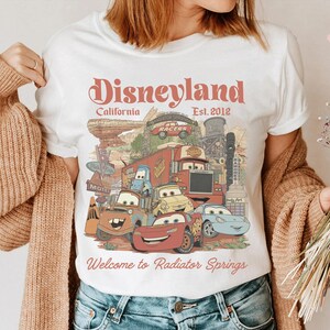 Peut inclure: T-shirt blanc avec un motif color&eacute; des personnages du film Cars, avec le texte "Disneyland California Est. 2012" et "Welcome to Radiator Springs". Le design comprend des voitures de dessins anim&eacute;s et des &eacute;l&eacute;ments de paysage.