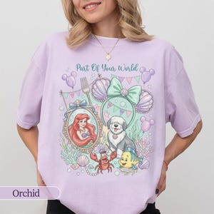 May include: A light purple t-shirt featuring a graphic print with the text Part Of Your World. The design includes a mermaid, a dog, a crab, a fish, seashells, bubbles, and mouse-ear headbands in pastel shades of pink, green, and purple.