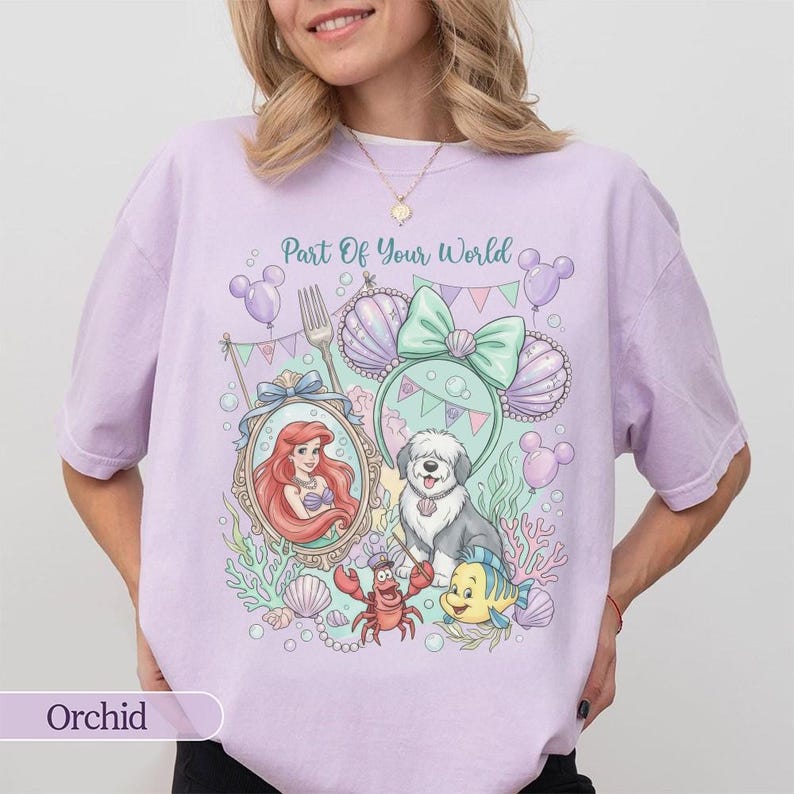 May include: A light purple t-shirt featuring a graphic print with the text Part Of Your World. The design includes a mermaid, a dog, a crab, a fish, seashells, bubbles, and mouse-ear headbands in pastel shades of pink, green, and purple.