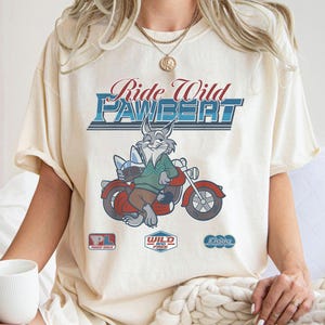 Puede incluir: Camiseta color crema con un conejo de dibujos animados montado en una motocicleta roja. La camiseta tiene el texto "Ride Wild Pawbert" en un estilo vintage, junto con logotipos m&aacute;s peque&ntilde;os. El conejo lleva un su&eacute;ter verde y pantalones marrones.