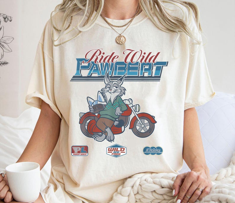 Puede incluir: Camiseta color crema con un conejo de dibujos animados montado en una motocicleta roja. La camiseta tiene el texto "Ride Wild Pawbert" en un estilo vintage, junto con logotipos m&aacute;s peque&ntilde;os. El conejo lleva un su&eacute;ter verde y pantalones marrones.