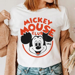 May include: White t-shirt featuring a Mickey Mouse Club design. The graphic includes Mickey Mouse's face, black ears, and the words "Mickey Mouse Club" in red, with a red circle around Mickey's face. The Disney logo is also visible.