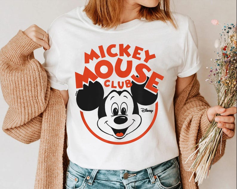 May include: White t-shirt featuring a Mickey Mouse Club design. The graphic includes Mickey Mouse's face, black ears, and the words "Mickey Mouse Club" in red, with a red circle around Mickey's face. The Disney logo is also visible.