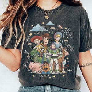 May include: A charcoal gray short-sleeve t-shirt featuring a graphic print of Toy Story characters including Woody, Buzz Lightyear, Jessie, Bo Peep, Rex, Hamm, Mr. Potato Head, Bullseye, and Forky, surrounded by stars, clouds, and handwritten names.