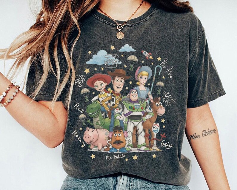 May include: A charcoal gray short-sleeve t-shirt featuring a graphic print of Toy Story characters including Woody, Buzz Lightyear, Jessie, Bo Peep, Rex, Hamm, Mr. Potato Head, Bullseye, and Forky, surrounded by stars, clouds, and handwritten names.